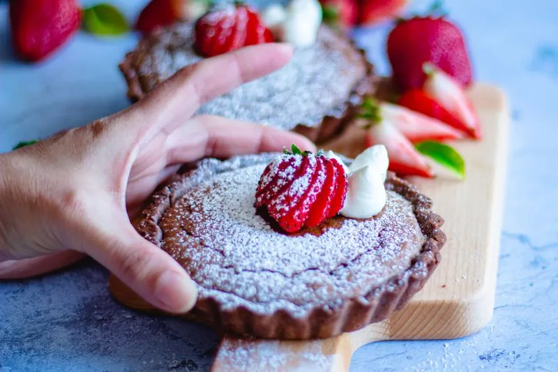 Chocolate tartlets dusted with icing sugar, topped with a strawberry and whipped cream, on a wooden board