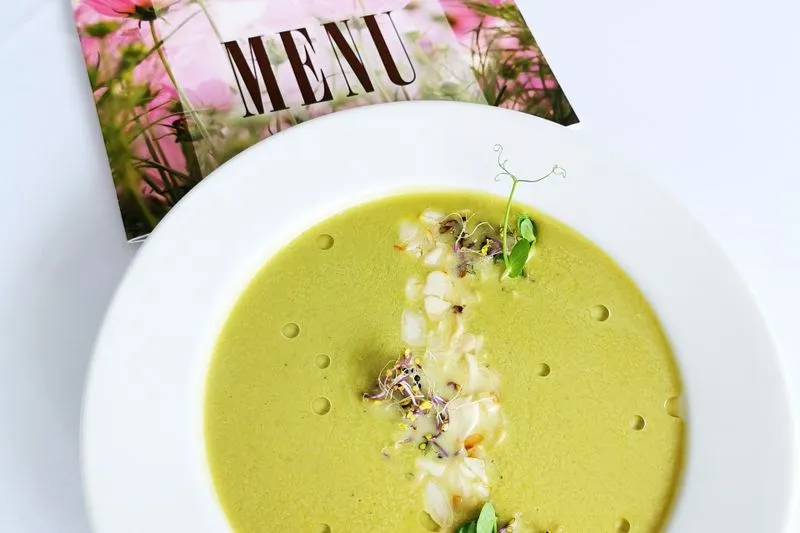 Pea soup in a white bowl, topped with pea shoots and sprouts, beside a menu card printed with flowers