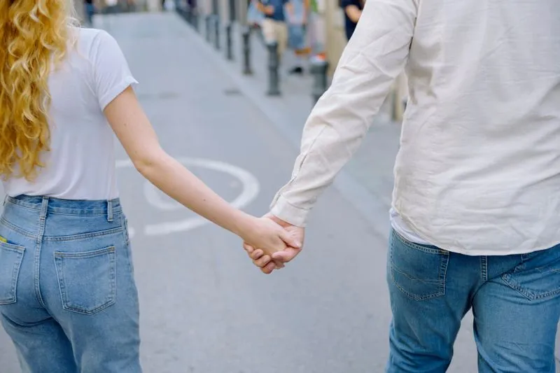 Couple seen from behind walking hand in hand down a pedestrian street, she in a white T-shirt, he in a linen shirt