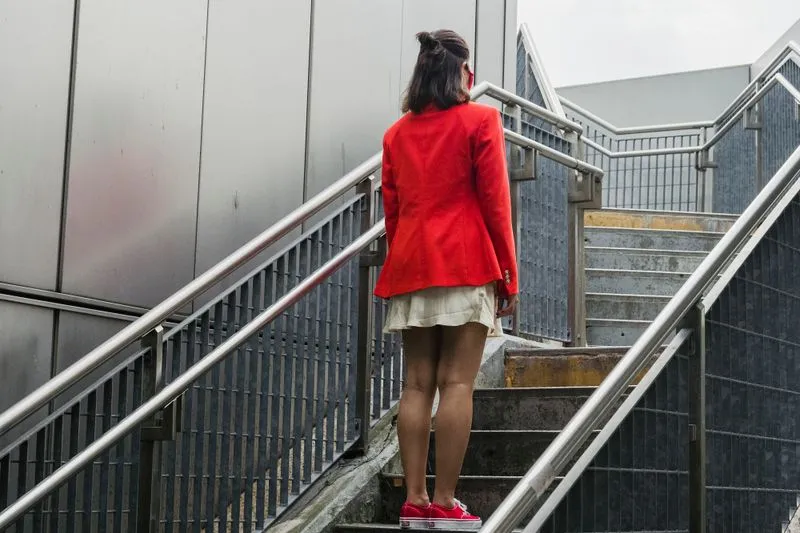 Young woman in a red blazer and red trainers, seen from behind, at the foot of a concrete staircase in town