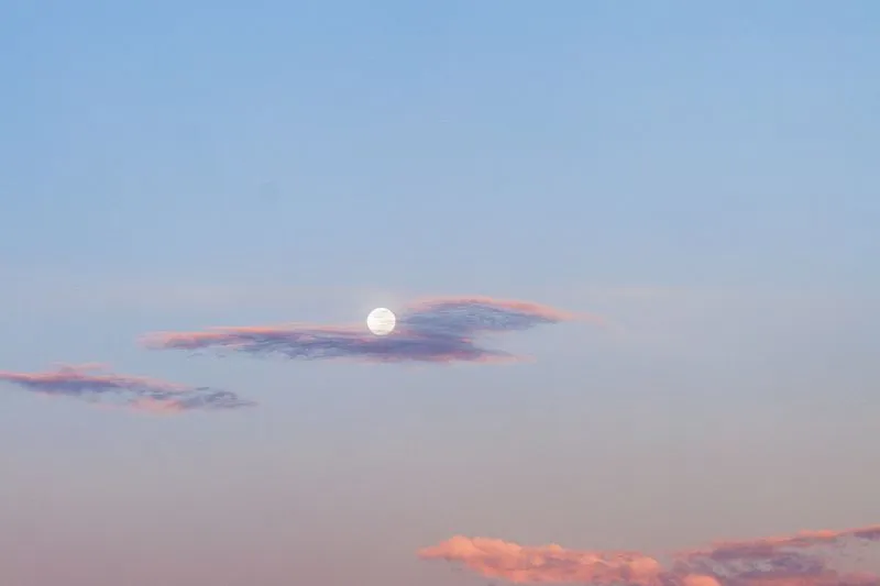 White full moon between pink-tinged clouds in a pale blue dusk sky