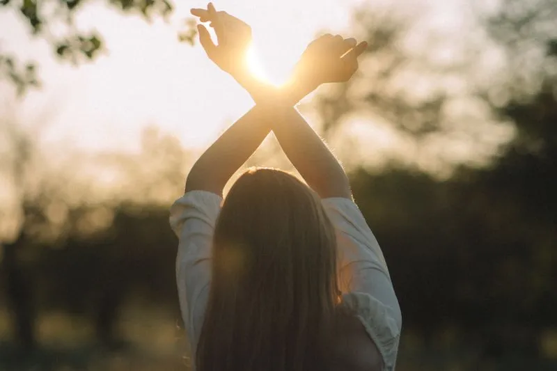 Young woman with long hair, seen from behind, arms raised and crossed towards low sunlight filtering through the trees