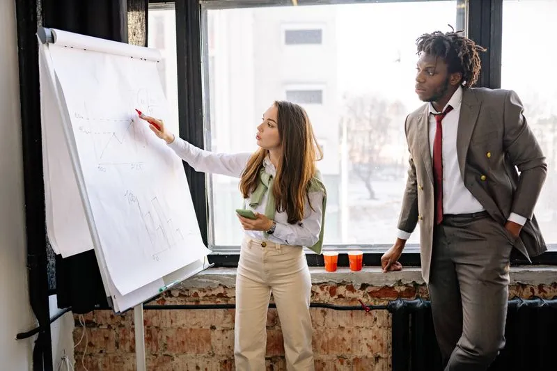 Young woman presenting graphs in red marker on a flip chart while a colleague in a suit listens closely