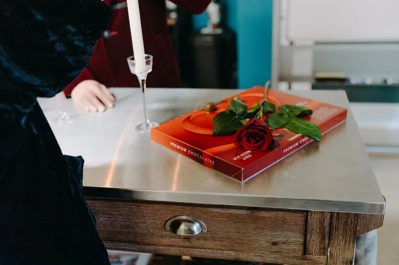 A red box of chocolates with a red rose laid across it, next to a candle holder