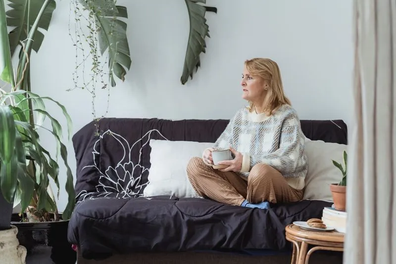 A woman in her fifties sitting cross-legged on a sofa, holding a mug in both hands