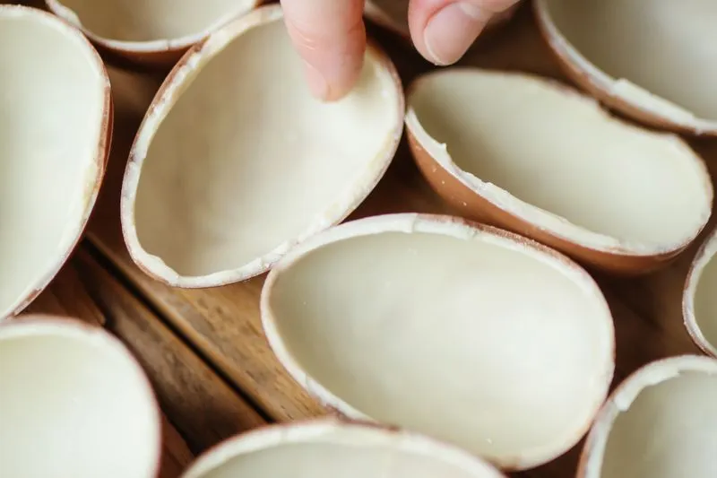 Hollow milk chocolate Easter egg halves laid out on a wooden table, one being set in place by hand