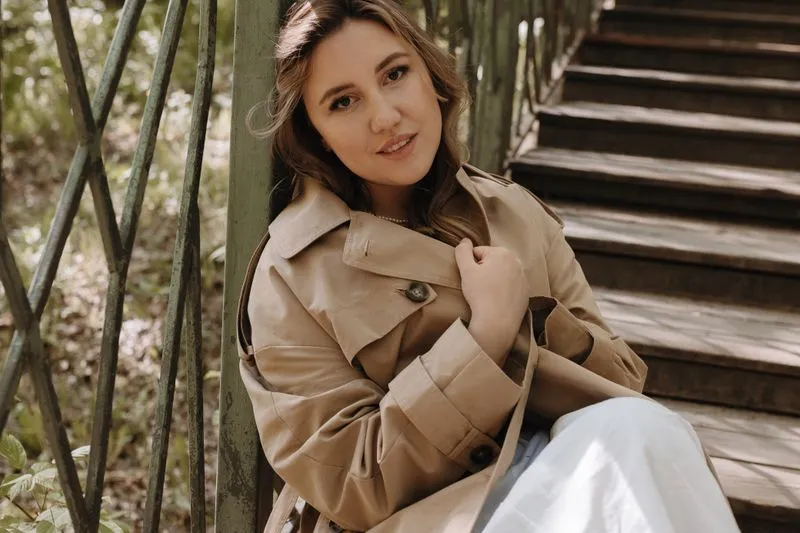 Woman sitting on wooden steps in a short beige trench coat: lapel collar, buttoned chest flap and cuff straps