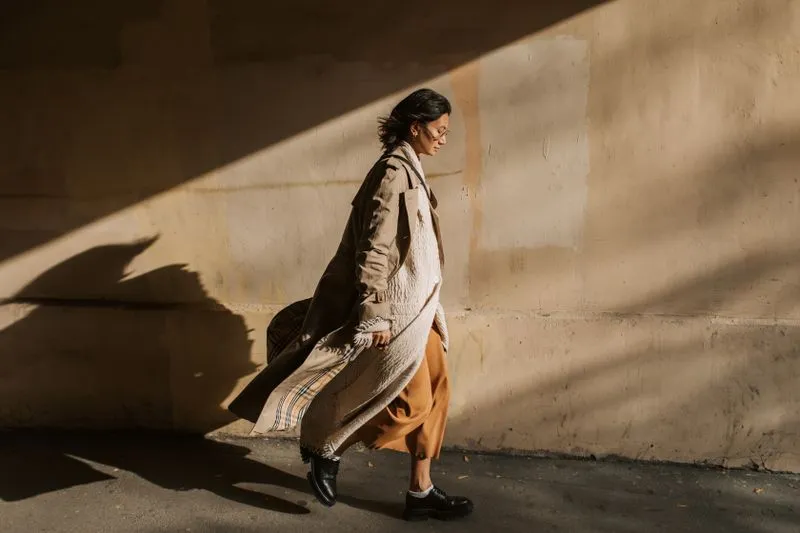 Woman walking past a sunlit wall in a long, open beige trench coat over a rust midi skirt and black derby shoes