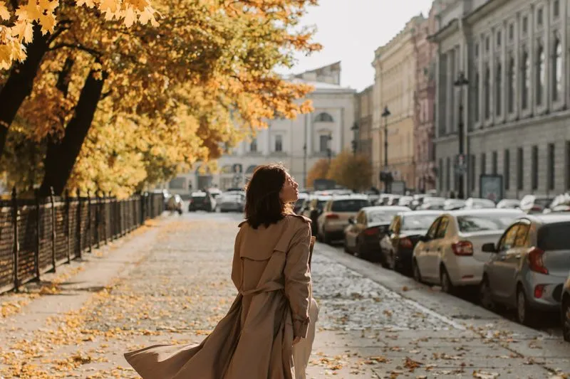 Woman seen from behind in a belted beige trench coat, its skirt lifted by the wind, on a cobbled avenue lined with old buildings and golden autumn trees