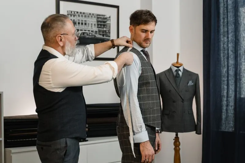 Tailor measuring a client's shoulder with a tape measure, a suit jacket on a mannequin behind them
