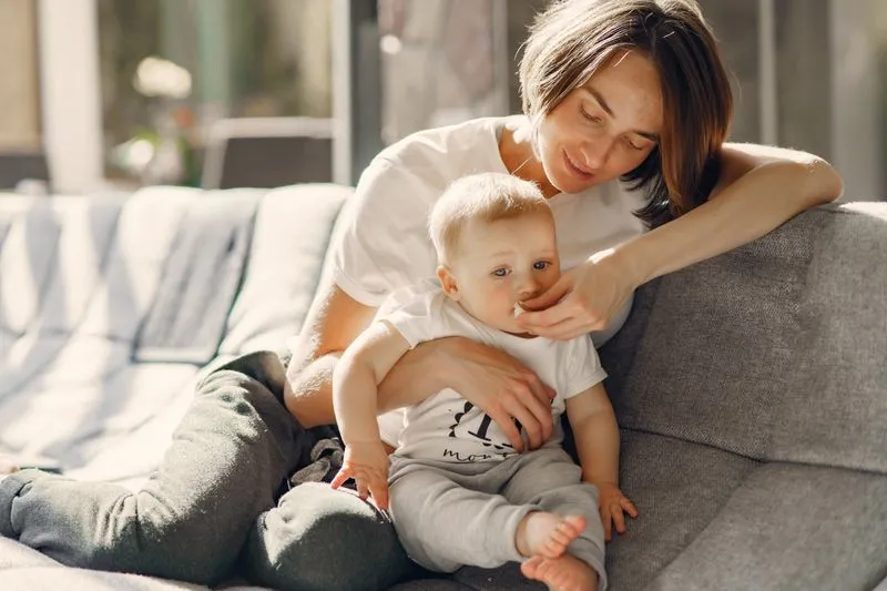 Young woman on a grey sofa with a baby on her lap, stroking his cheek in a sunlit living room
