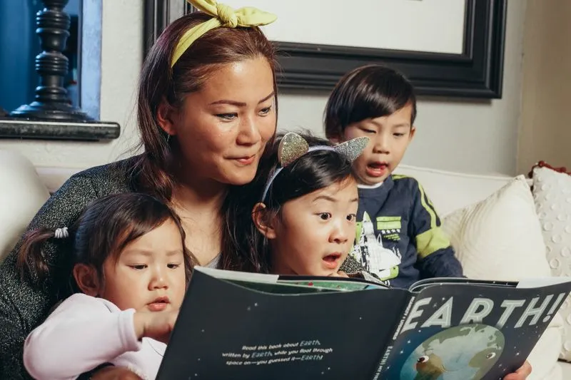 Woman on a sofa reading a picture book to three young children huddled around her