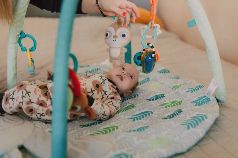 Baby lying on a play mat under a baby gym, smiling up at hanging toys that an adult's hand is moving