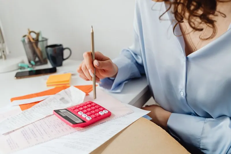 Woman in a pale blue blouse holding a pencil over a pink calculator resting on bills and receipts