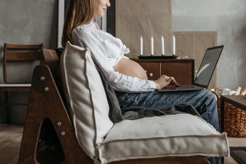 Pregnant woman sitting in a wooden armchair typing on a laptop on her lap