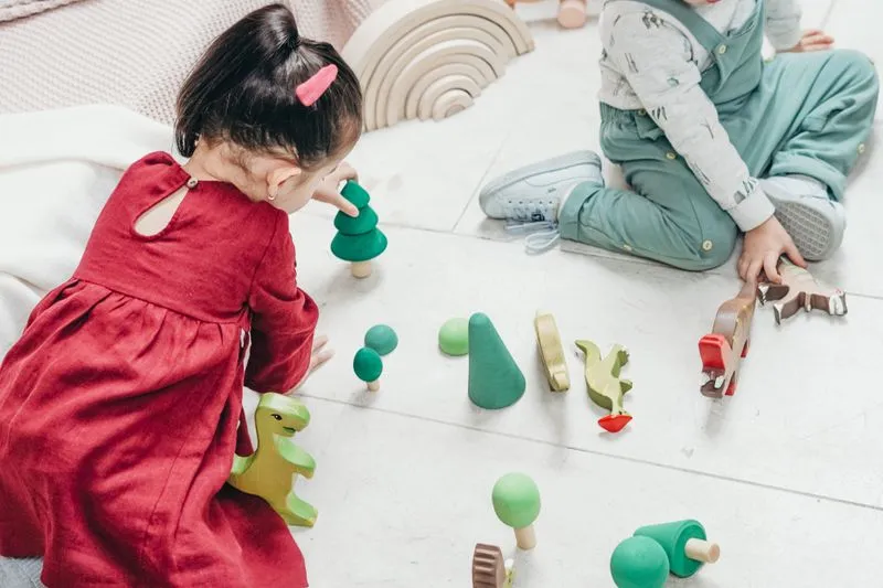 Two toddlers sitting on the floor playing with wooden trees and dinosaurs, a stacking rainbow behind them