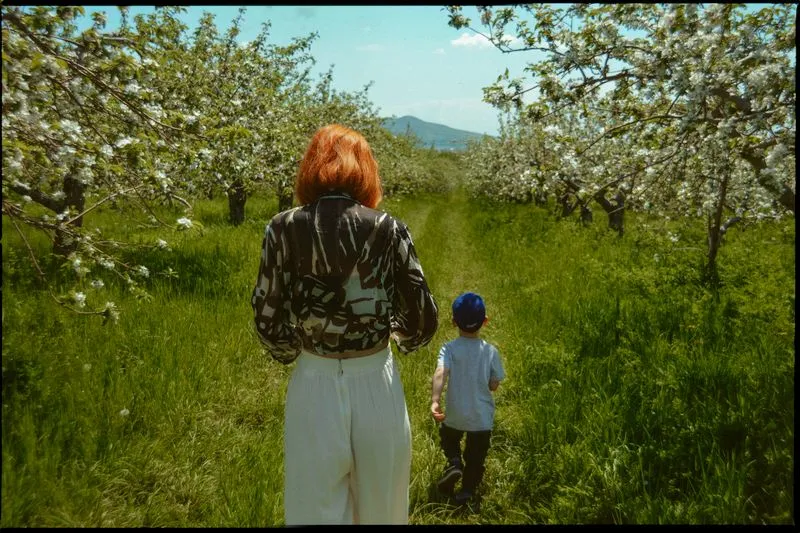 Red-haired woman walking behind a little boy in a cap through an apple orchard in blossom