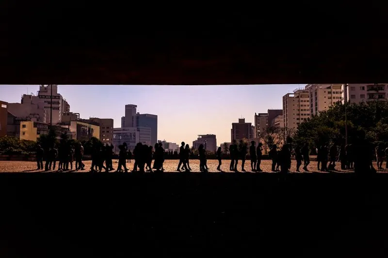 Silhouettes of a long queue of people waiting beneath a large concrete overhang, with tower blocks behind