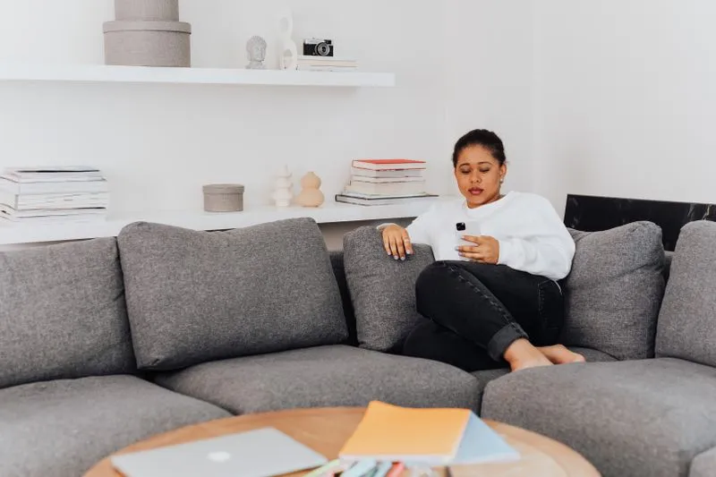 Young woman in a white jumper sitting on a grey sofa, looking at her smartphone