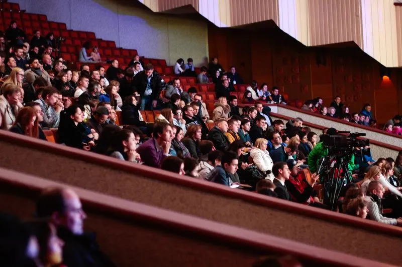 Audience seated in the red tiered rows of a large hall, with a camera operator filming from among the seats