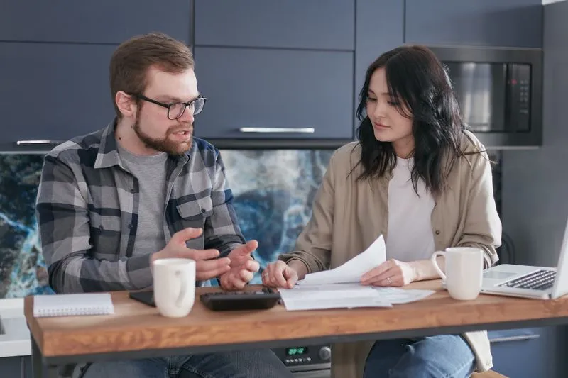 Couple sitting at a kitchen counter, he explains with his hands while she holds papers; calculator, mugs and a laptop in front of them