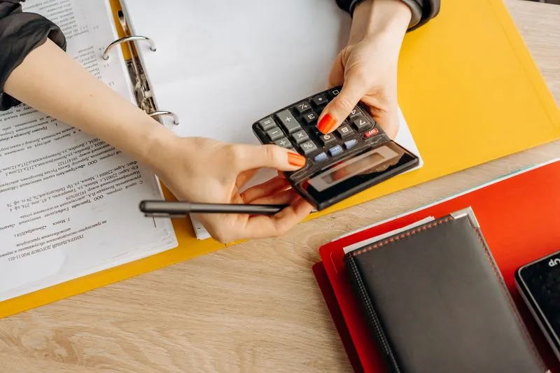 A woman's hands holding a calculator and a pen over an open binder full of documents