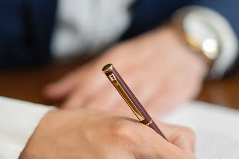 Hand signing a document with a burgundy and gold pen, a second person blurred in the background