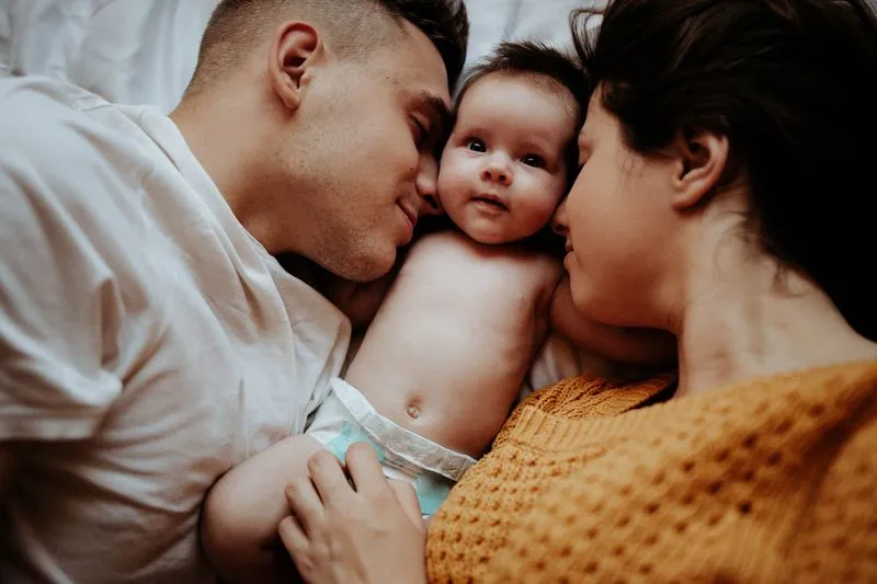 Young couple lying on a bed, cheek to cheek with their baby lying between them