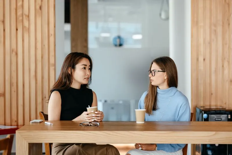 Two young women talking across a large wooden table, each with a paper coffee cup