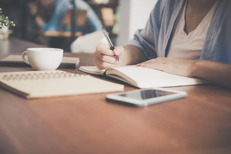 A woman's hand writing in an open notebook on a wooden table, next to a white cup and a phone