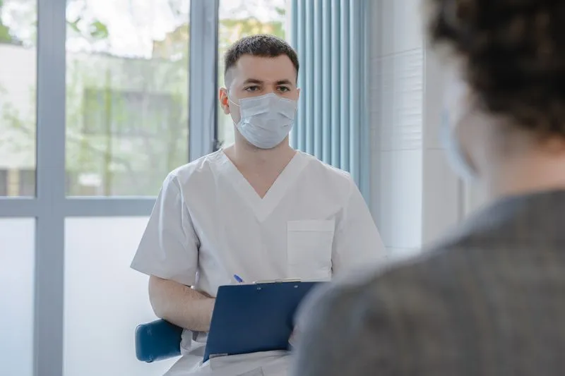 A masked clinician in a white tunic taking notes on a clipboard, facing a patient seen from behind