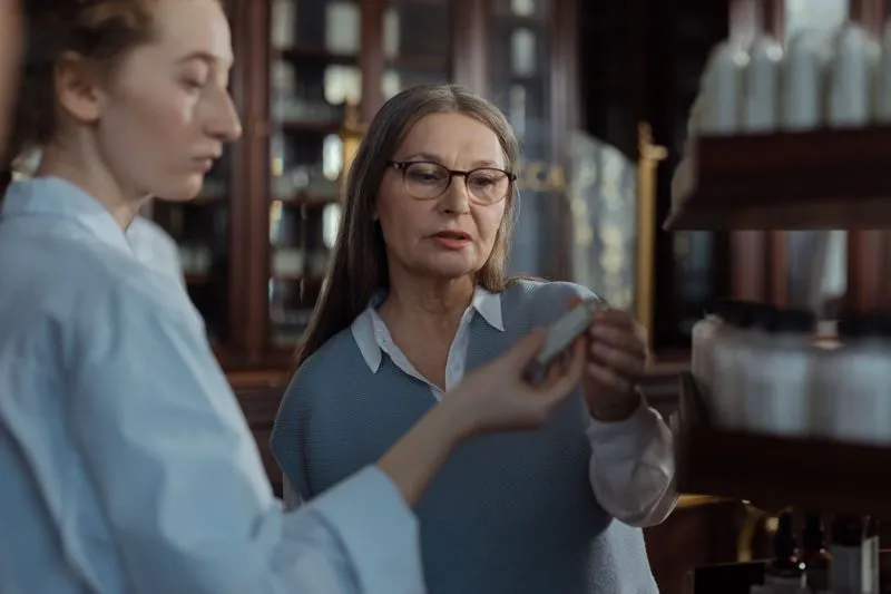 A young woman in a white shirt showing a small bottle to a woman in glasses, in front of wooden shelves lined with bottles