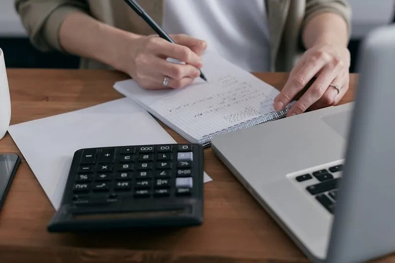 A hand noting calculations in a spiral notebook, next to a calculator and a laptop
