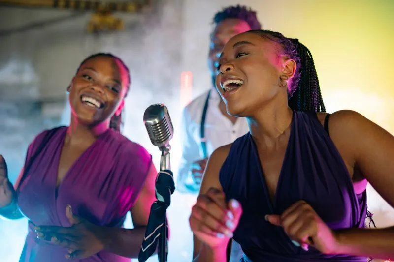 Two young women laughing as they sing into a vintage microphone under pink and purple lights