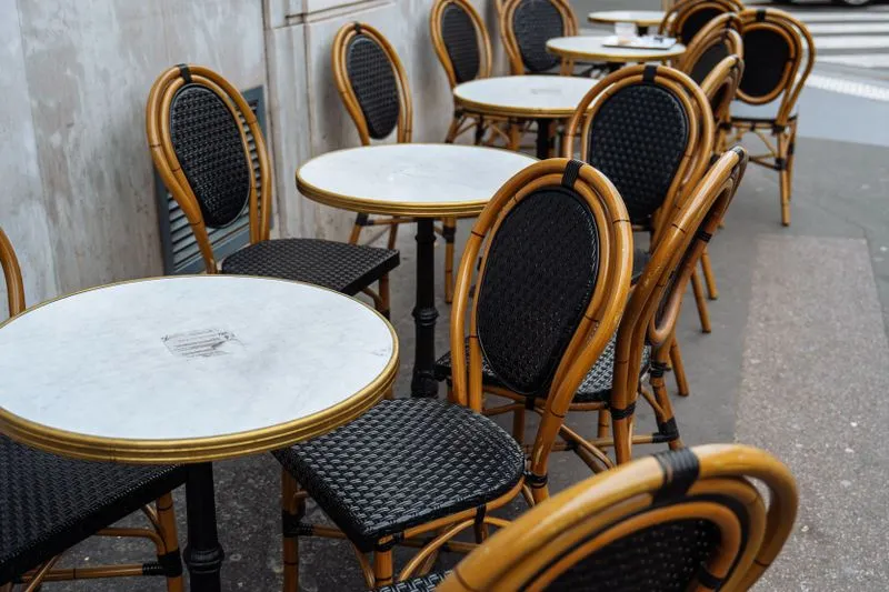 Round marble tables edged in brass and woven rattan bistro chairs lined up on the pavement of a Paris café terrace