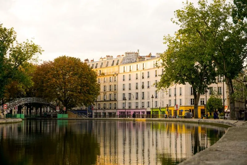 Canal Saint-Martin quay in late-afternoon light, an iron footbridge on the left and pale buildings reflected in the water