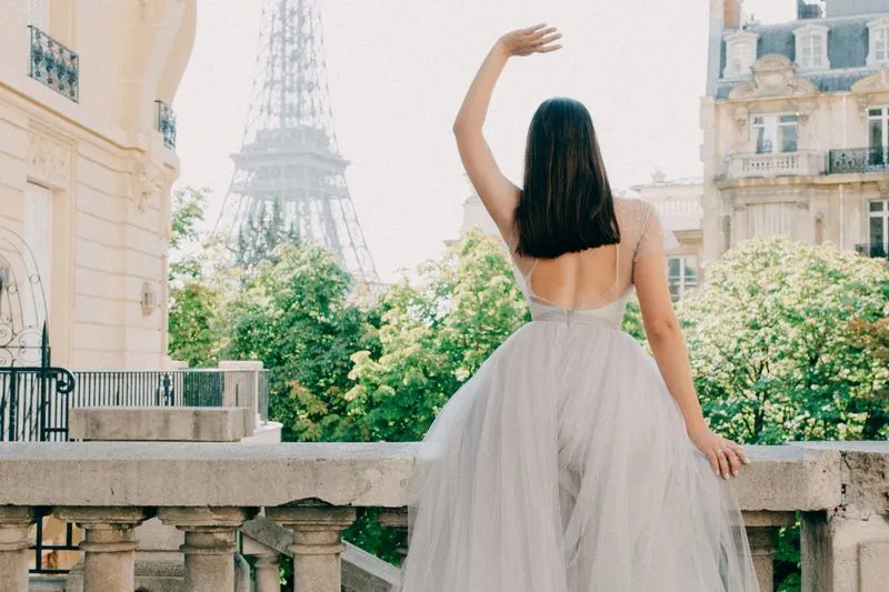 Young woman in a grey tulle dress seen from behind on a Paris stone balcony, one arm raised towards the Eiffel Tower