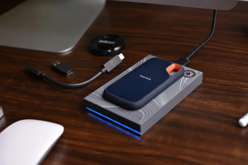 Portable SSD resting on an external hard drive, with its USB-C cable, on a dark wooden desk