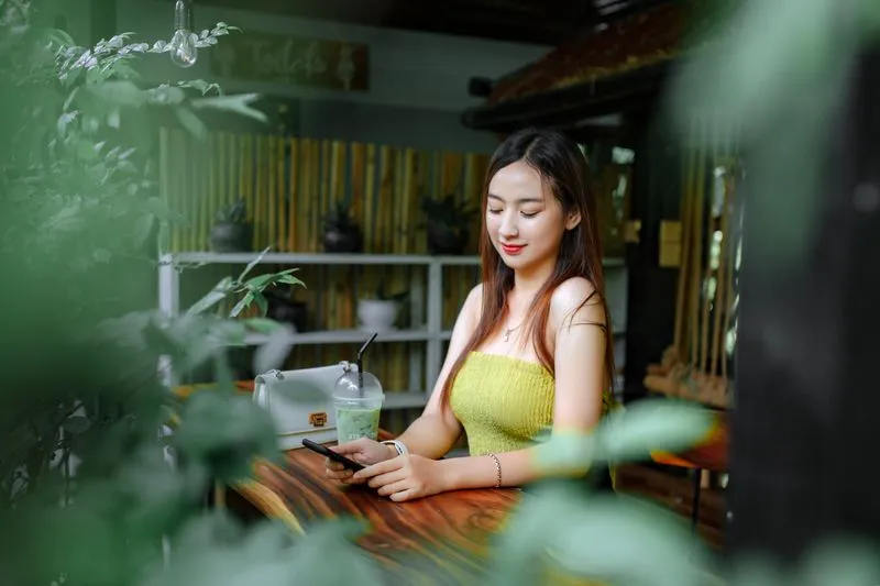 Young woman at a café table surrounded by plants, looking at her smartphone, an iced drink beside her