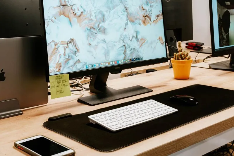 Wooden desk with a monitor, a laptop closed on its stand and a smartphone in front of the keyboard