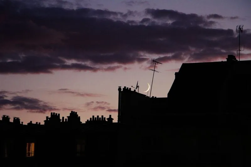 Thin crescent moon between the chimneys and aerials of city rooftops at dusk