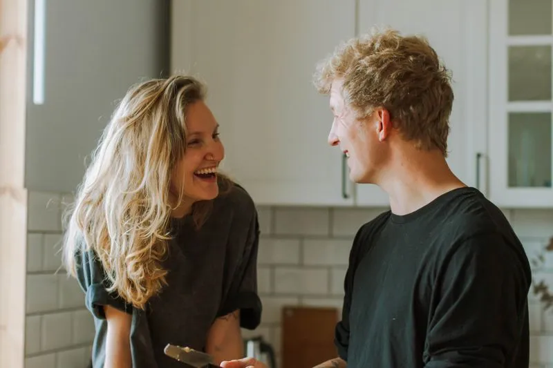 A woman and a man laughing face to face in their kitchen, one of them holding a knife