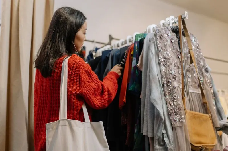 Young woman in a red jumper, canvas bag on her shoulder, looking through the clothes on a charity shop rail