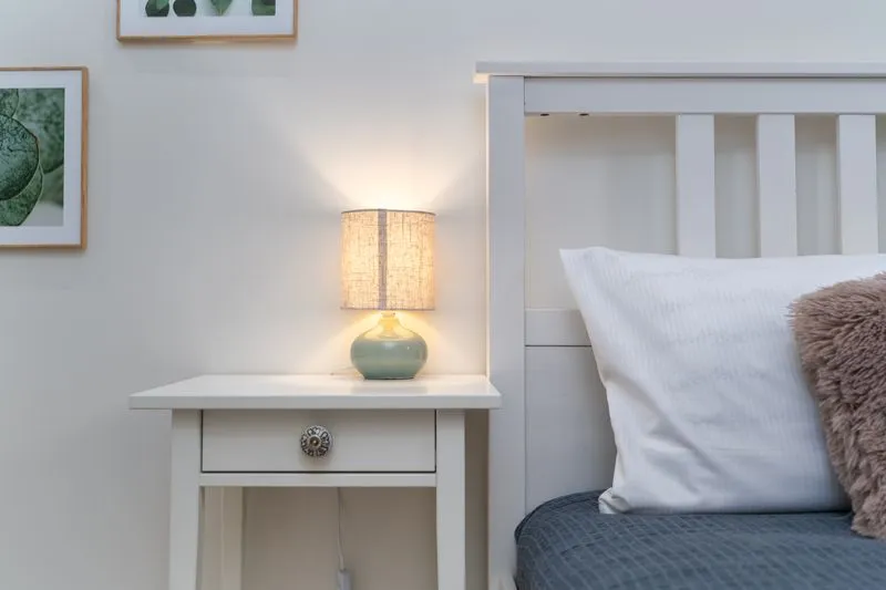 Bedside lamp switched on, on a white nightstand beside a bed and its pillow
