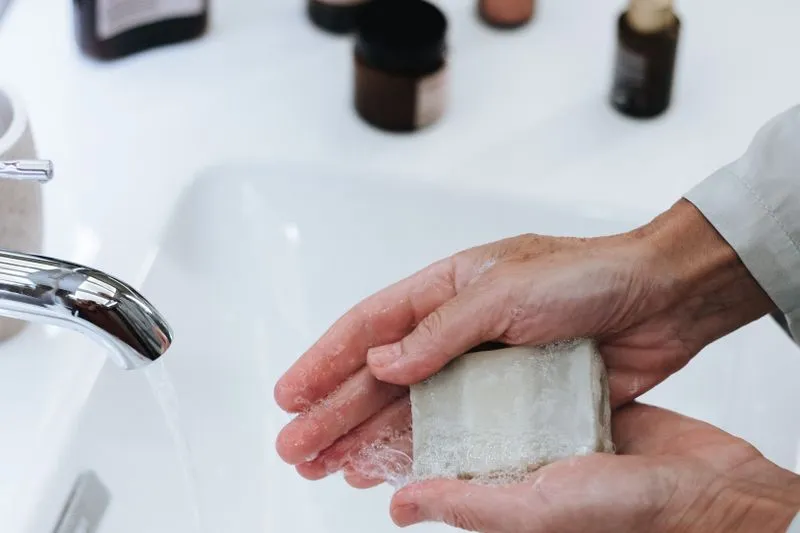 Hands lathering a bar of soap under a running tap, above a white basin