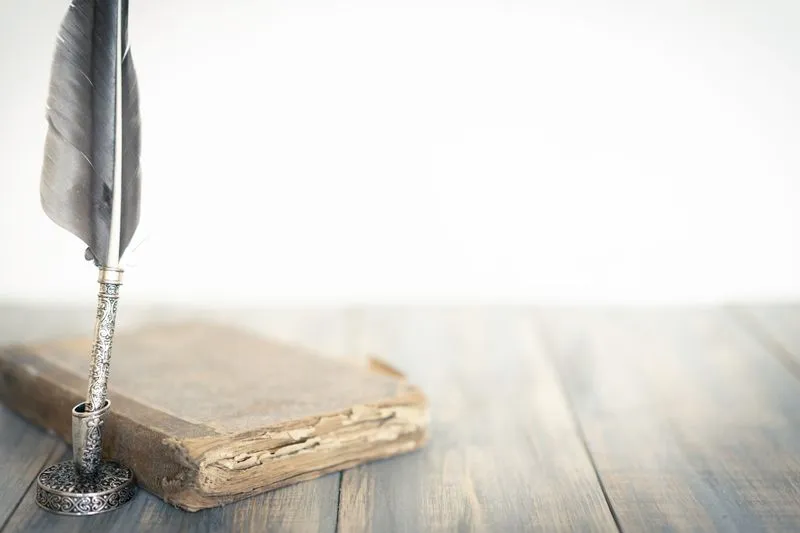 Old bound book with a worn cover on a wooden table, next to a quill pen