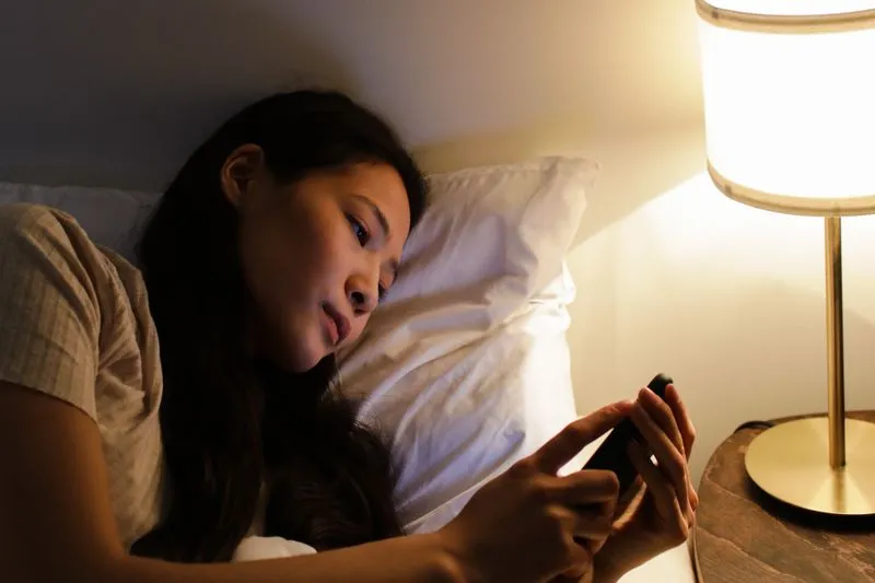Young woman lying in bed in the evening, eyes on her phone, beside a lit bedside lamp
