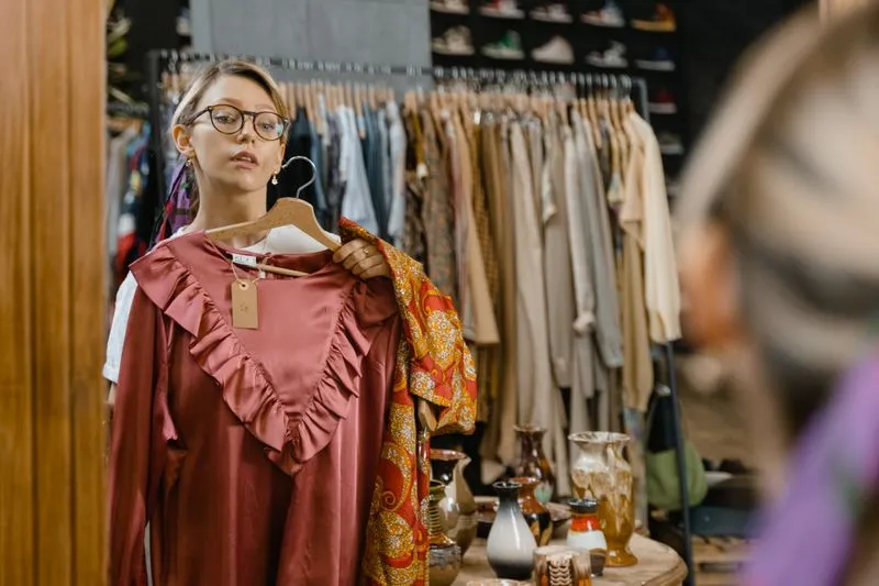 Young woman in glasses holding a plain dusty-rose blouse against herself in a vintage shop mirror