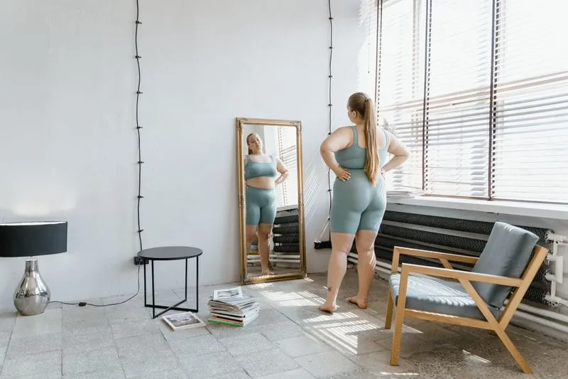 Woman standing in front of a large gilt mirror propped against the wall, next to a sunny window