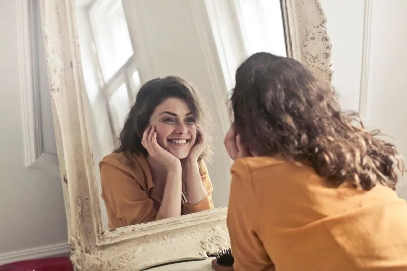 Young woman in a mustard top smiling at herself in a large antique mirror near a window